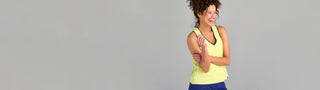 A woman with curly hair smiles while wearing The Jenni - Ace tank top, with blue shorts, standing against a plain gray background.