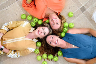Overhead view of moorlow’s three female founders lying with heads together, surrounded by green pickleballs—playful, purposeful portrait of the women shaping pickleball style.