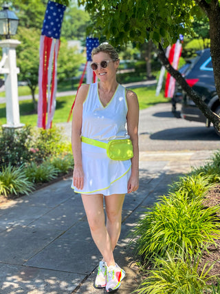 Woman in a white moorlow tank top and skort  with a green fanny pack walking on a sidewalk with American flags in the background.