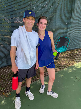 Two people on a pickleball court, one holding a paddle and a ball wearing a moorlow tank top and skort, the other wearing a blue moorlow cap.