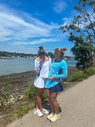 Two women standing on a path by a body of water with a clear blue sky wearing moorlow athletic pullovers and skorts.