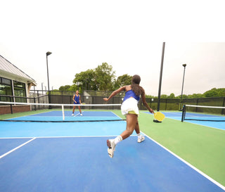 Two women playing pickleball on a blue and green court with trees in the background.